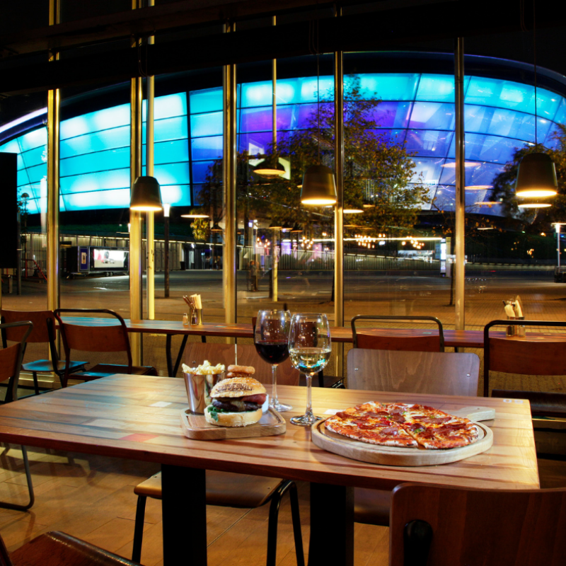 Inside clydebuilt with a burger, pizza, red wine, and white wine sitting on a table with the OVO Hydro in the background