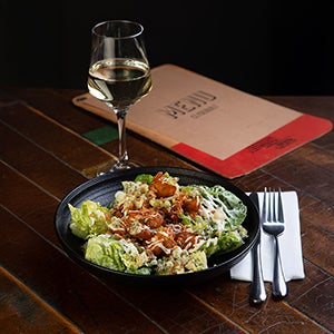 Bowl of salad on a table with cutlery, a glass of wine and menu next to it. 