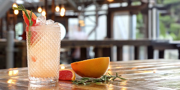 A table in Clydebuilt with a highball glass on top of it which has ice, drink, a bit of herb and a grapefruit segment in it. The remaining grapefruit and herb is also on the table.